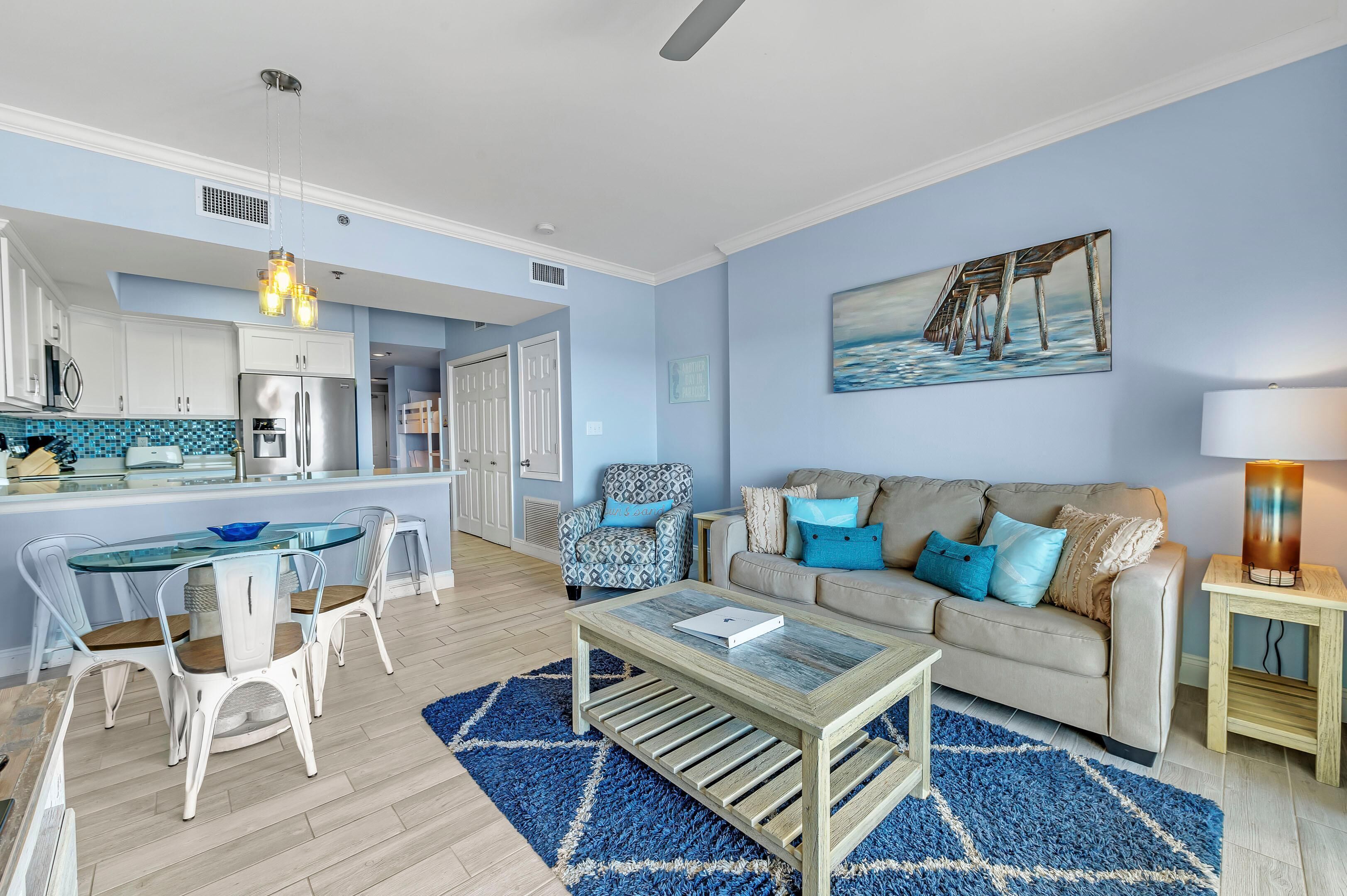 A modern living room with a glass dining table, a beige sofa with blue cushions, a blue rug, and a painting of a pier on the wall.