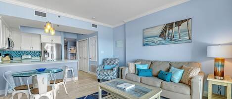 A modern living room with a glass dining table, a beige sofa with blue cushions, a blue rug, and a painting of a pier on the wall.