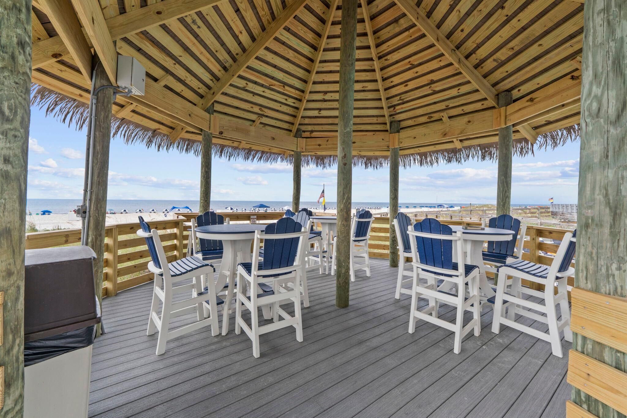 Un espacio de estar al aire libre techado con mesas y sillas de madera, con vista a una playa.