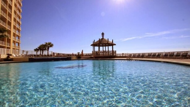 A clear pool with a gazebo in the center, surrounded by a high-rise building and palm trees. - Summer House 1501b (Orange Beach)