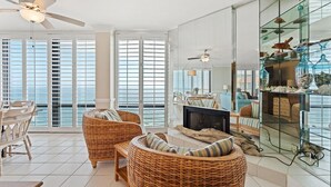 A spacious living area with wicker furniture, a glass coffee table, and a television. Large windows offer a view of the ocean. - Summer House 1501b (Orange Beach)