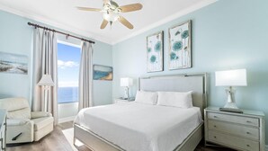 A bedroom with a bed, a chair, a nightstand, a lamp, and a ceiling fan. - Spanish Key 702 (Pensacola)