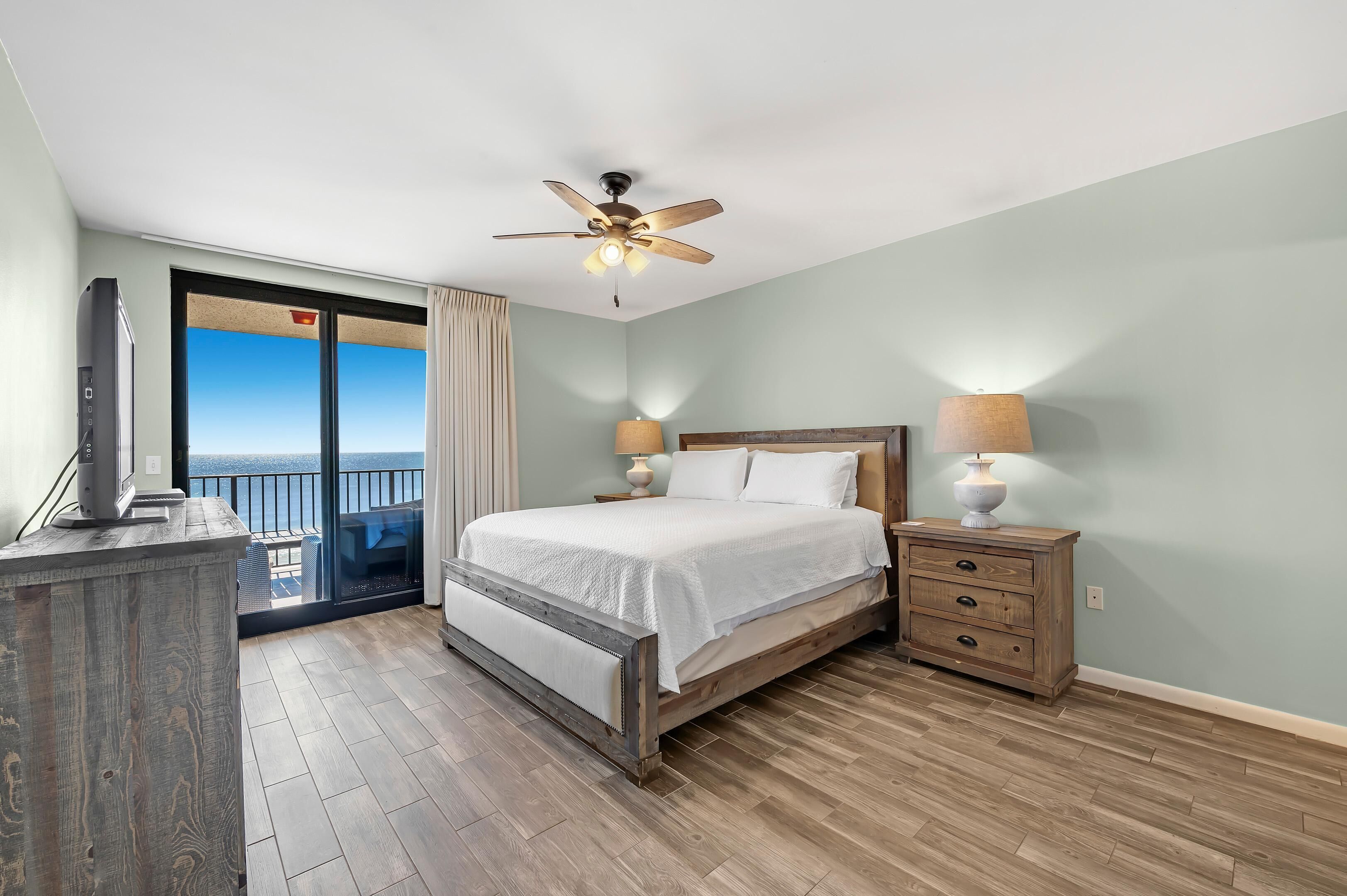 A bedroom with a bed, bedside lamps, a ceiling fan, and a view of the outdoors.