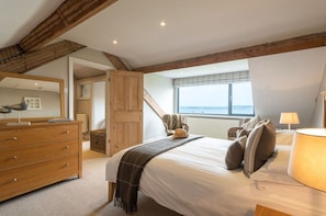 A bedroom with a large bed, wooden beams, a dresser, a chair, and a window with a view. - Seascape: A fabulous, pet-friendly home with sea views (Rhosneigr)