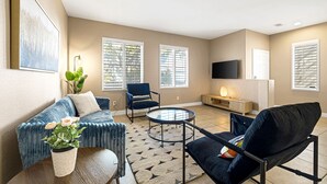 Una sala de estar moderna con sofá, sillón, mesa de centro, televisor de pantalla plana y ventanas amplias con persianas.