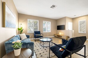 Een moderne woonkamer met een bank, fauteuil en salontafel, een flatscreen-televisie en grote ramen met zonwering.