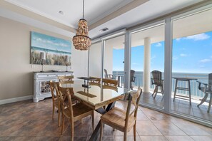 A dining area with a glass table and wooden chairs, a chandelier, a painting, and a view of the ocean through large sliding glass doors.