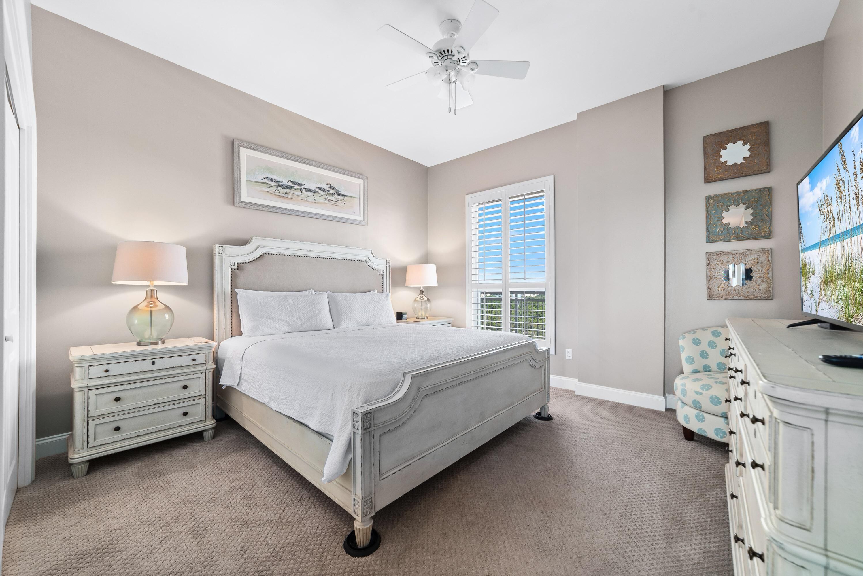 A bedroom with a bed, two nightstands, a ceiling fan, a window with blinds, a TV, and a dresser.