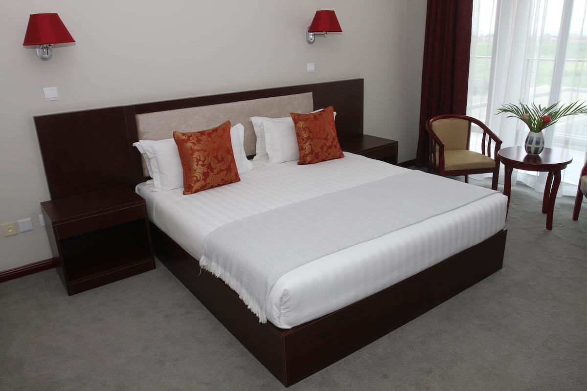 Una cama grande con sábanas blancas y dos almohadas rojas, una mesita de noche de madera, una silla y una pequeña mesa con un jarrón de flores.