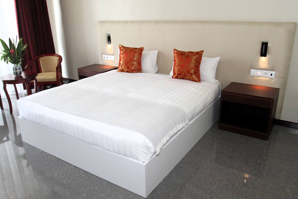 Una cama grande con sábanas blancas, dos almohadas rojas y dos mesitas de noche con lámparas fijadas a la pared.
