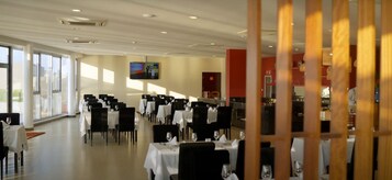 Un comedor espacioso con mesas dispuestas para varios comensales, ventanas amplias que dejan entrar luz natural y un televisor de pantalla plana montado en la pared.