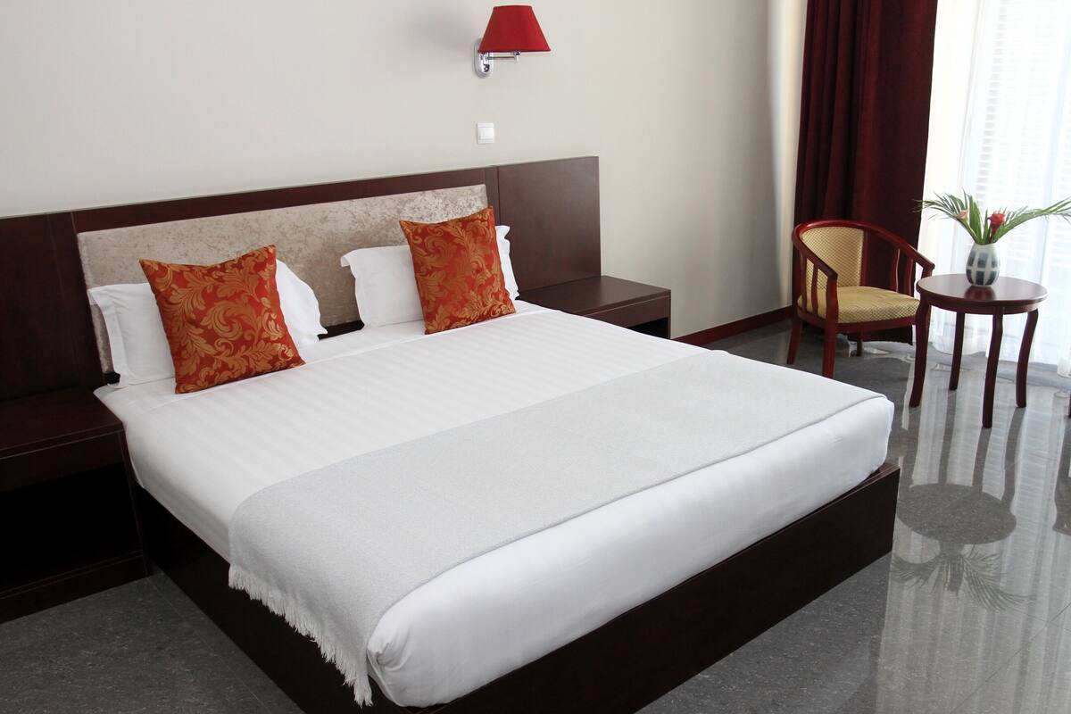 Una habitación de hotel con una cama grande, dos mesitas de noche, una lámpara de pared roja, una silla, una pequeña mesa y un florero con flores.