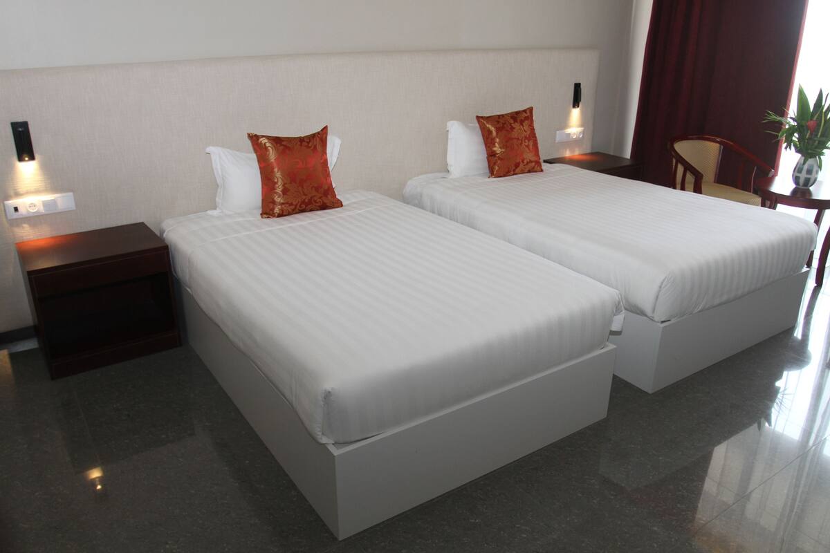 Habitación de hotel con dos camas, cada una con ropa blanca y almohadas decorativas rojas. Hay mesitas de noche con lámparas, una silla y una pequeña mesa con una planta.