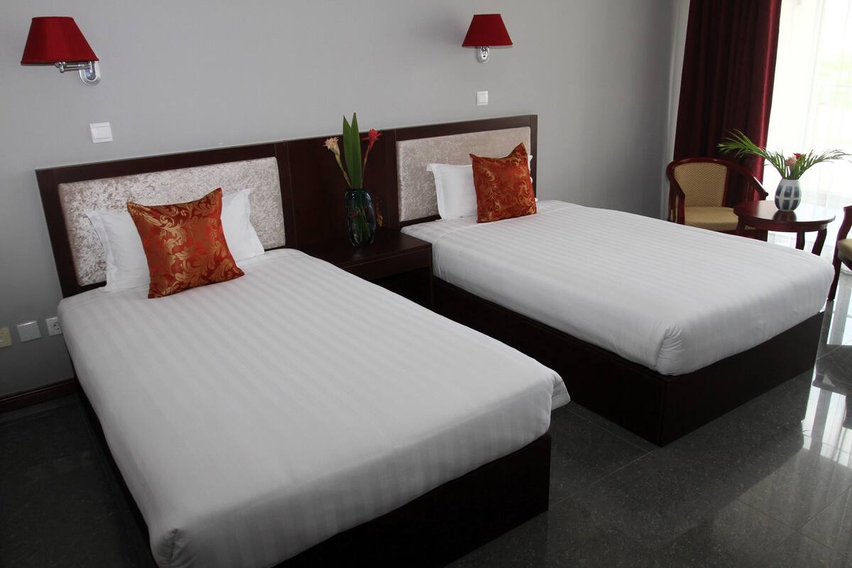 Dos camas individuales con ropa blanca y almohadones rojos, una mesita de noche con un florero, una silla y una pequeña mesa con una planta.