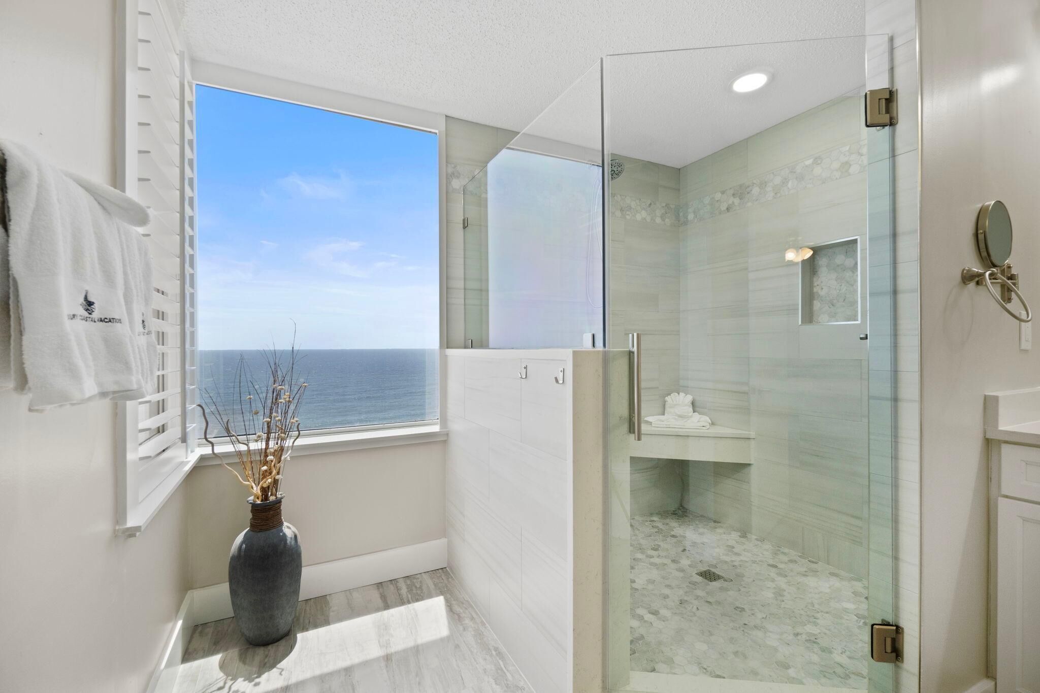 Un baño moderno con ducha envuelta en vidrio, un ventanal con vista al mar y un florero con ramas secas en el alféizar.