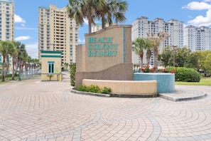 A sign for Beach Colony Resort with a paved area, palm trees, and high-rise buildings in the background. - Beach Colony West 11B (Pensacola)