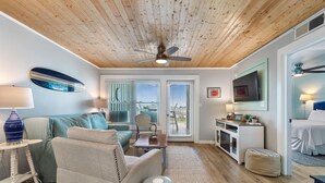 A living room with a wooden ceiling, a sofa, a coffee table, a TV, and a door leading outside. - Jubilee Landing 206 (Orange Beach)
