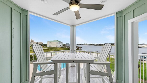 A covered outdoor area with a table and chairs, a ceiling fan, and a view of a body of water and buildings in the distance. - Jubilee Landing 206 (Orange Beach)