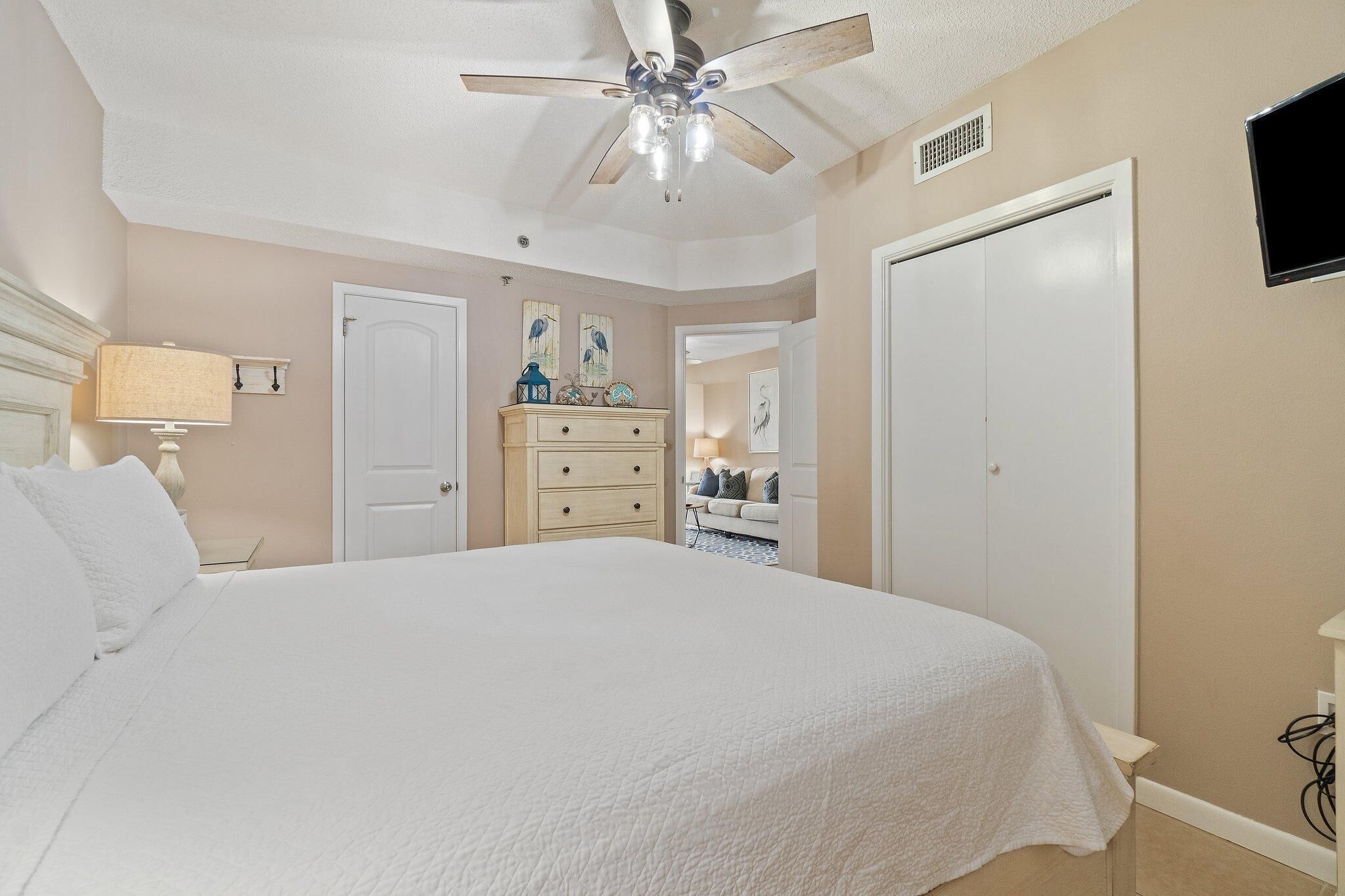 A bedroom with a large bed, a ceiling fan, a dresser, a television, and a door.