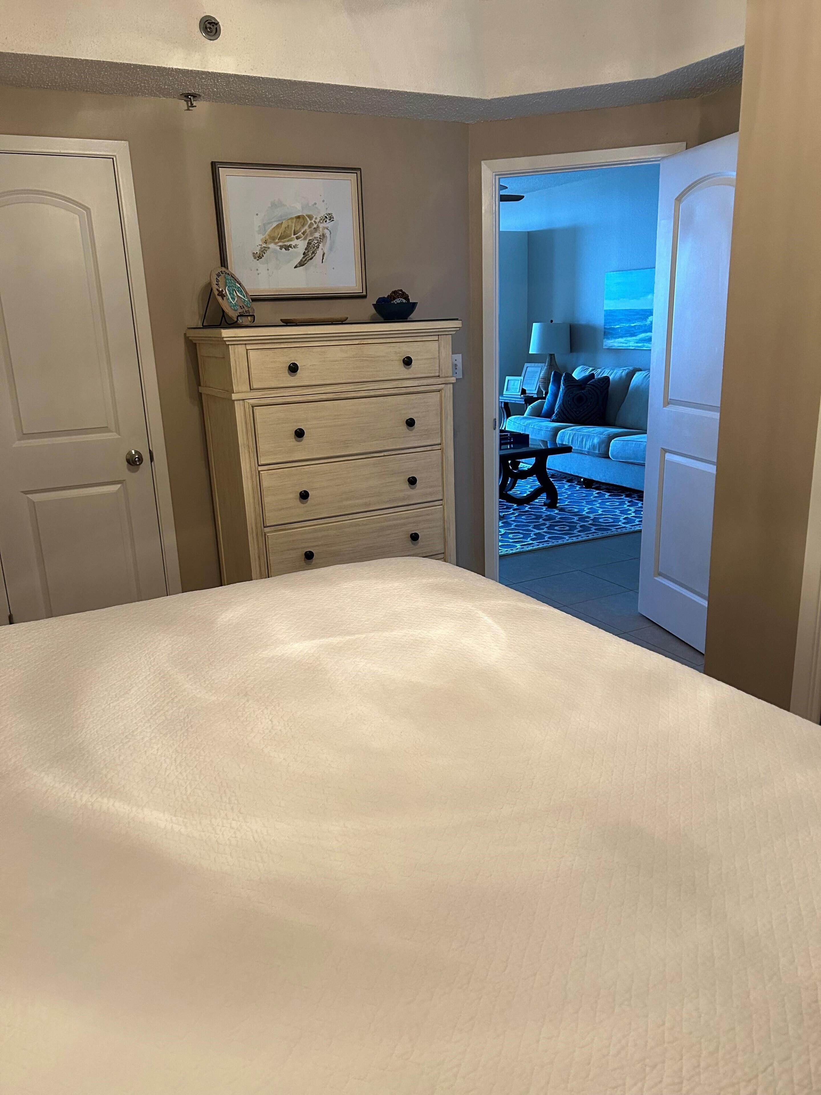 A bedroom with a bed, a dresser, and a door leading to a living area.