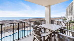 Un balcon avec vue sur une piscine et une plage, des meubles en bois et, au loin, un immeuble de grande hauteur.