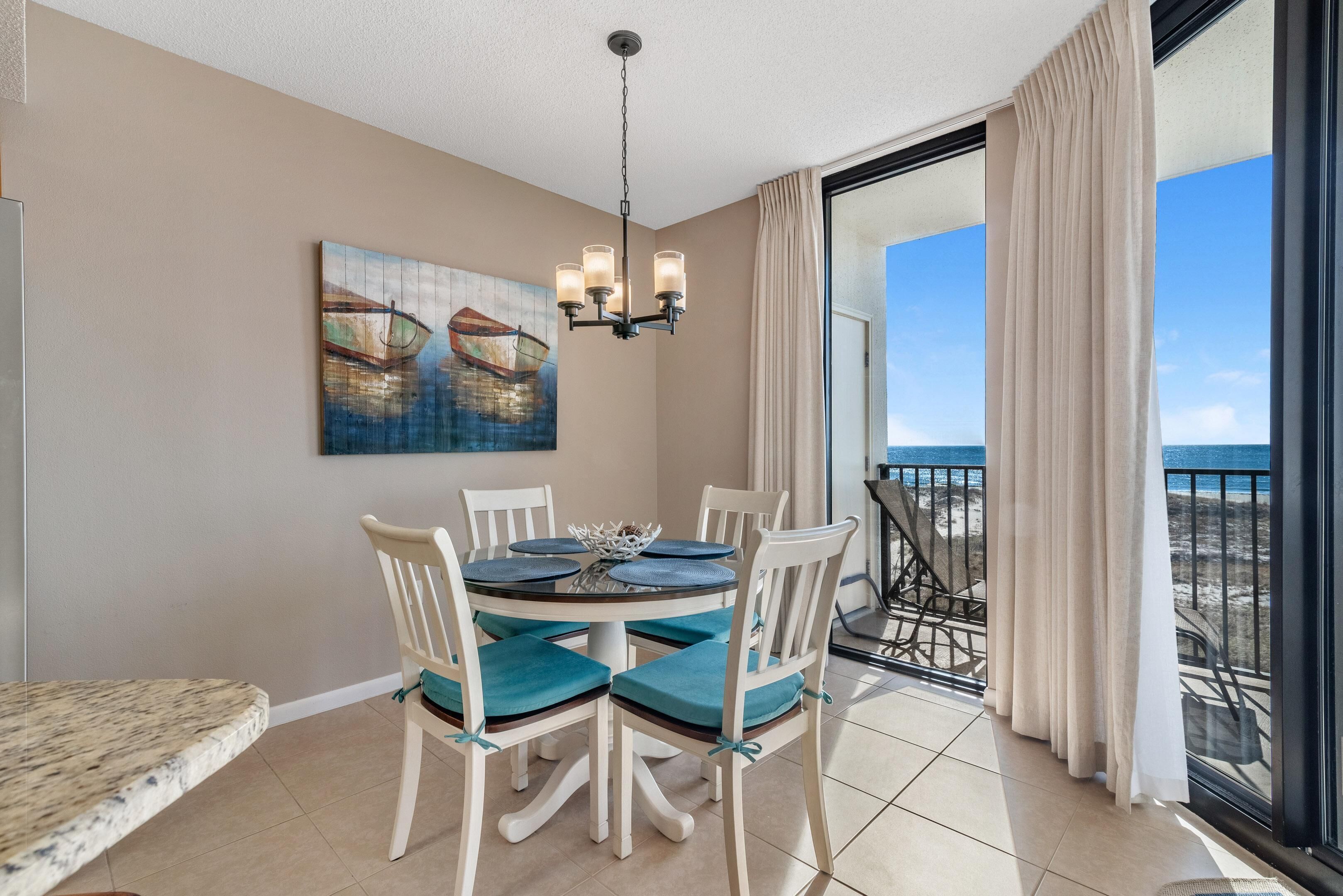 Un coin repas avec une table ronde, des chaises blanches et des coussins turquoise. Un lustre est suspendu au plafond, et un tableau représentant des bateaux est accroché au mur.