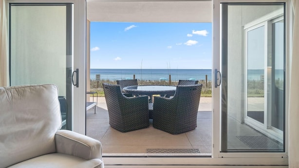 Une porte-fenêtre coulissante donne accès à un espace extérieur aménagé avec une table et des chaises, et offre une vue sur la plage.