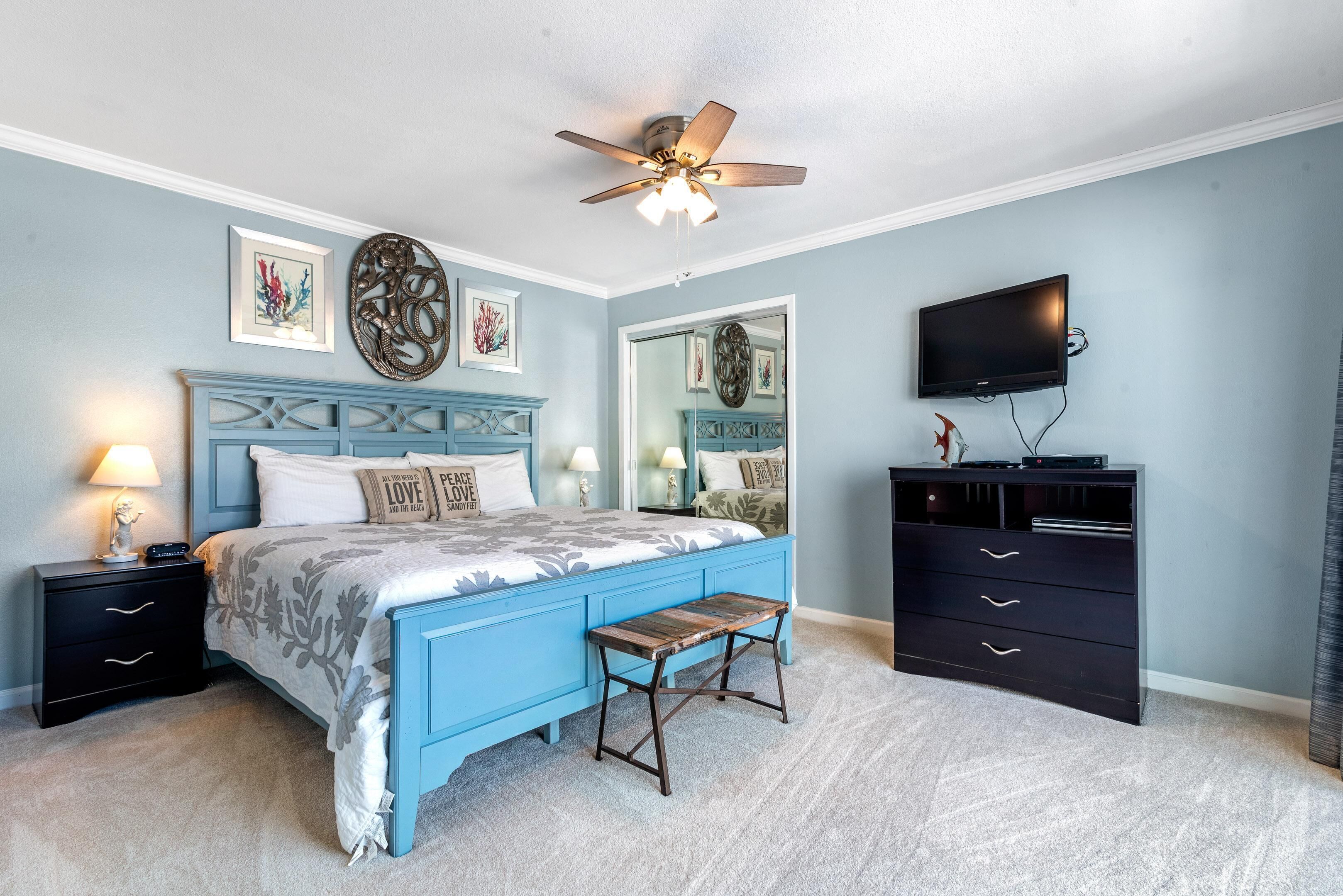A bedroom with a blue bed, a wooden bench, a ceiling fan, a TV, and a dresser.