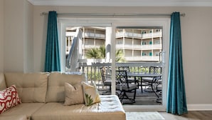 A living room with a beige sofa, teal curtains, and a view of a balcony with outdoor furniture. - Boardwalk F3 (Pensacola Beach)