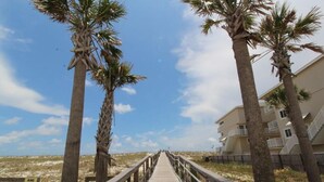A wooden boardwalk leading to a beach, flanked by palm trees and a building with balconies. - Boardwalk F3 (Pensacola Beach)
