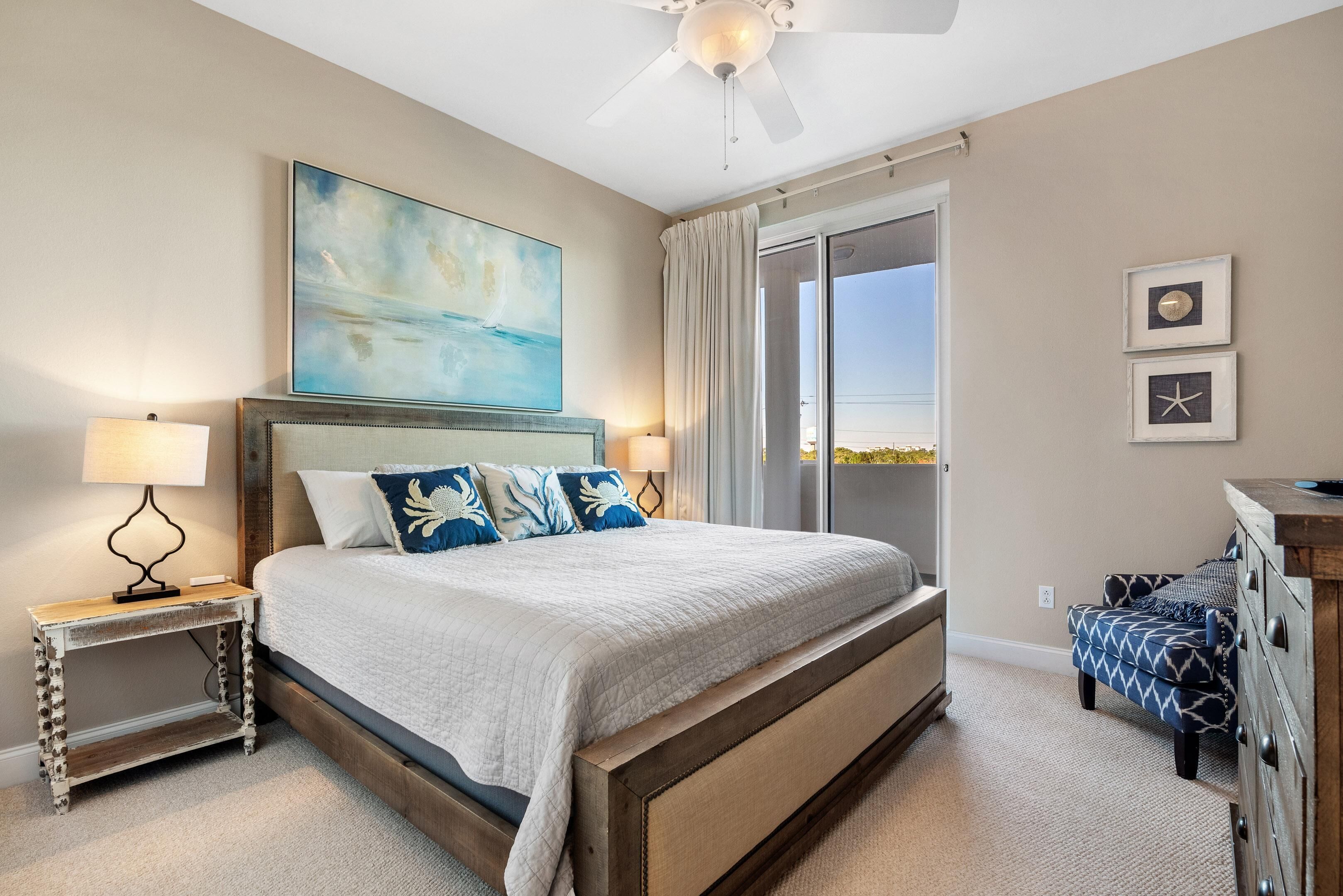 A bedroom with a large bed, a ceiling fan, a painting, and a view of the outdoors through a sliding door.