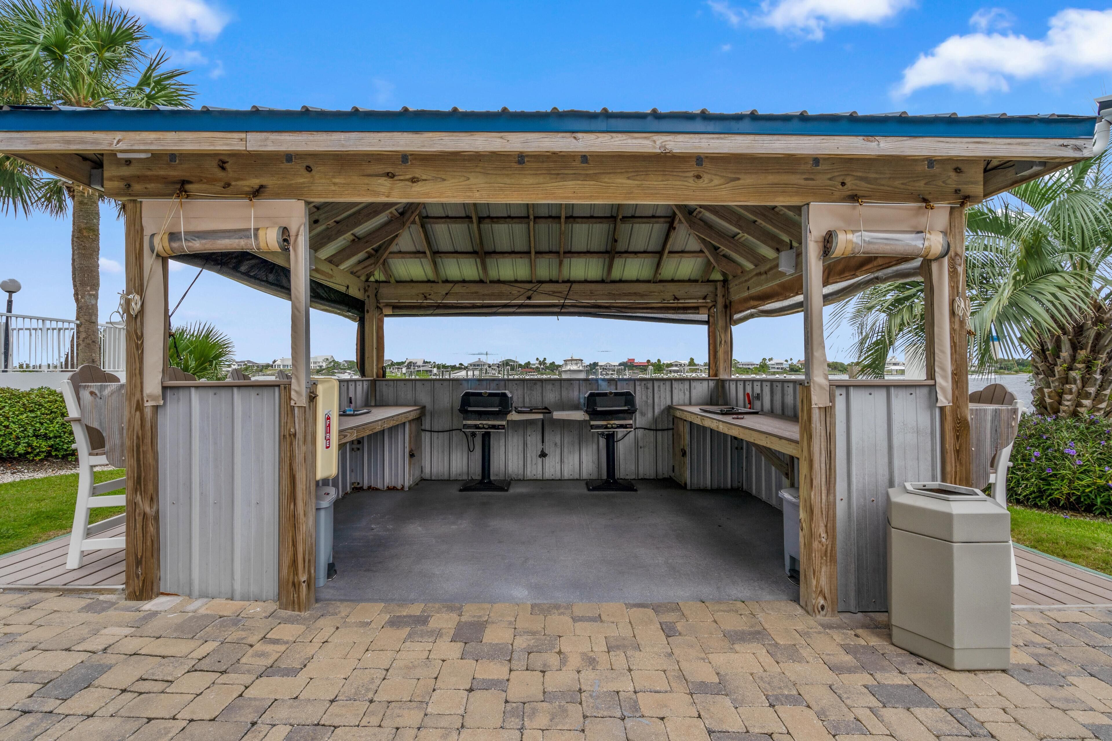 Zona de barbacoa al aire libre con un pérgola de madera, dos parrillas y un bote de basura.