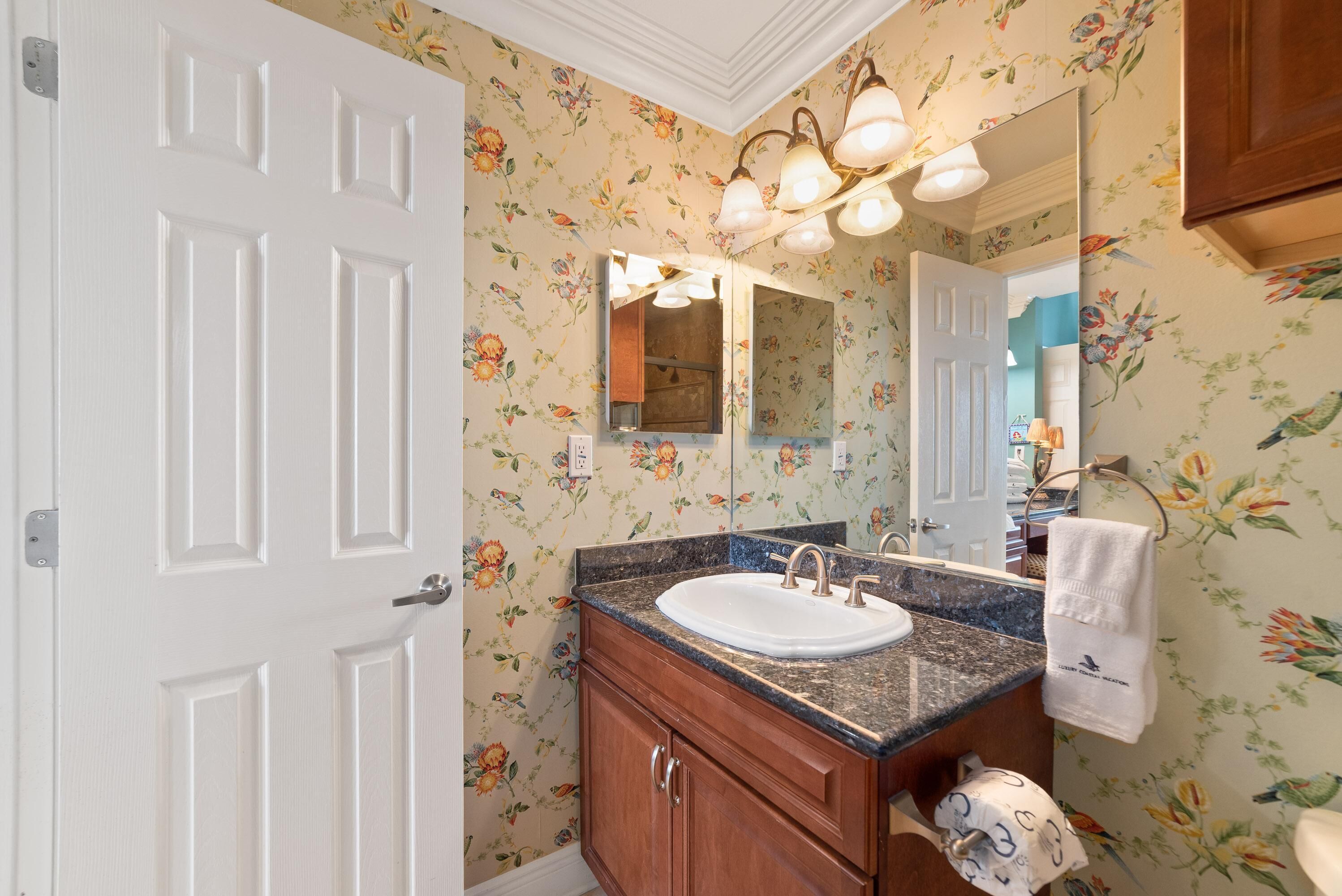 Une salle de bain avec un papier peint à motifs floraux, un lavabo blanc, des meubles en bois et un miroir avec éclairage intégré.