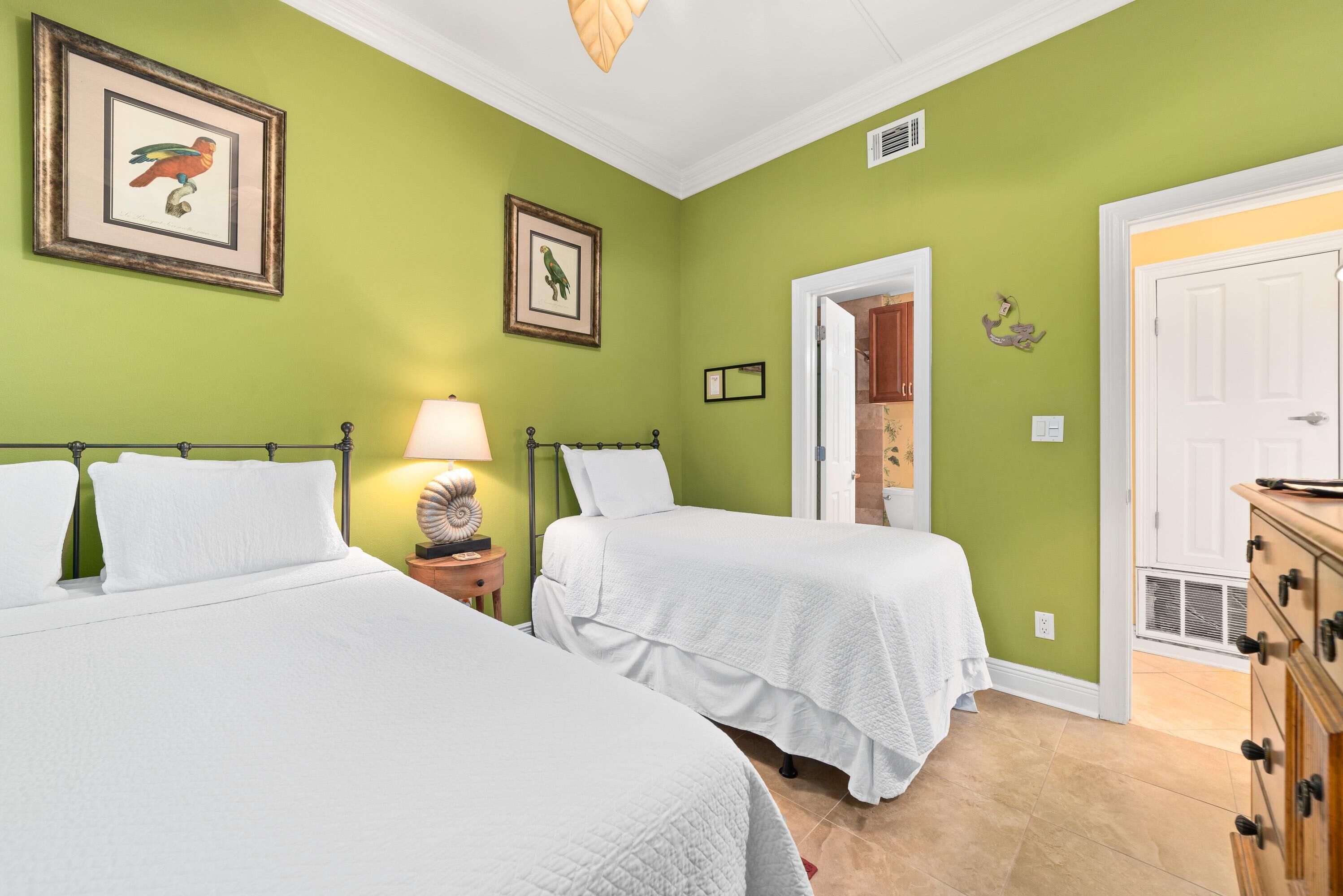 A room with two beds, green walls, framed pictures, a lamp, and a wooden dresser.