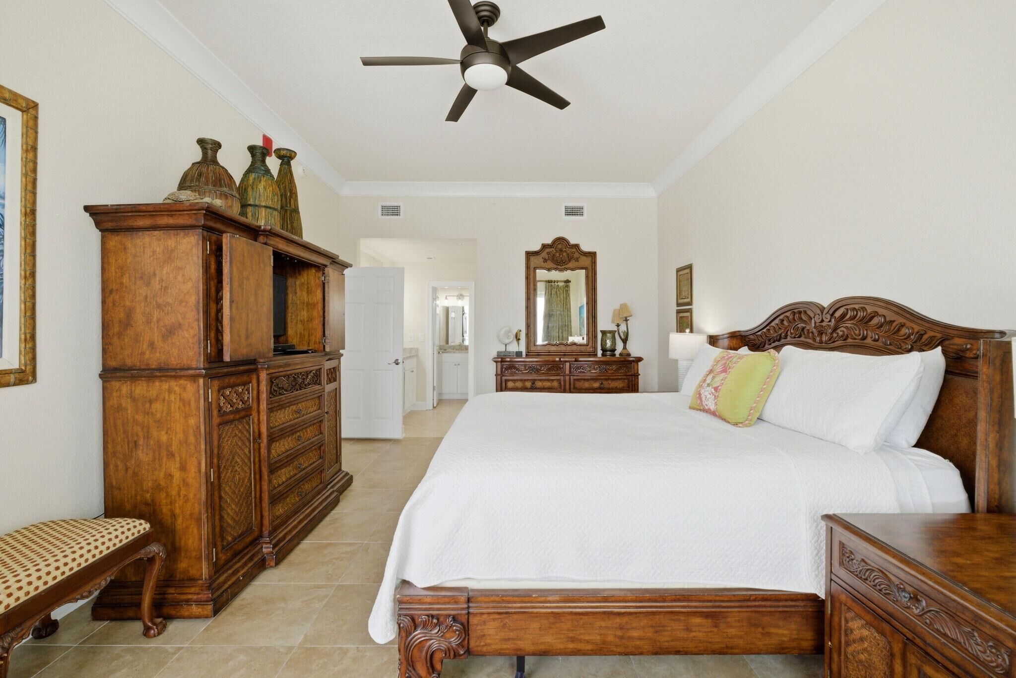 A bedroom with a large bed, a wooden dresser, a bench, a ceiling fan, and a mirror.