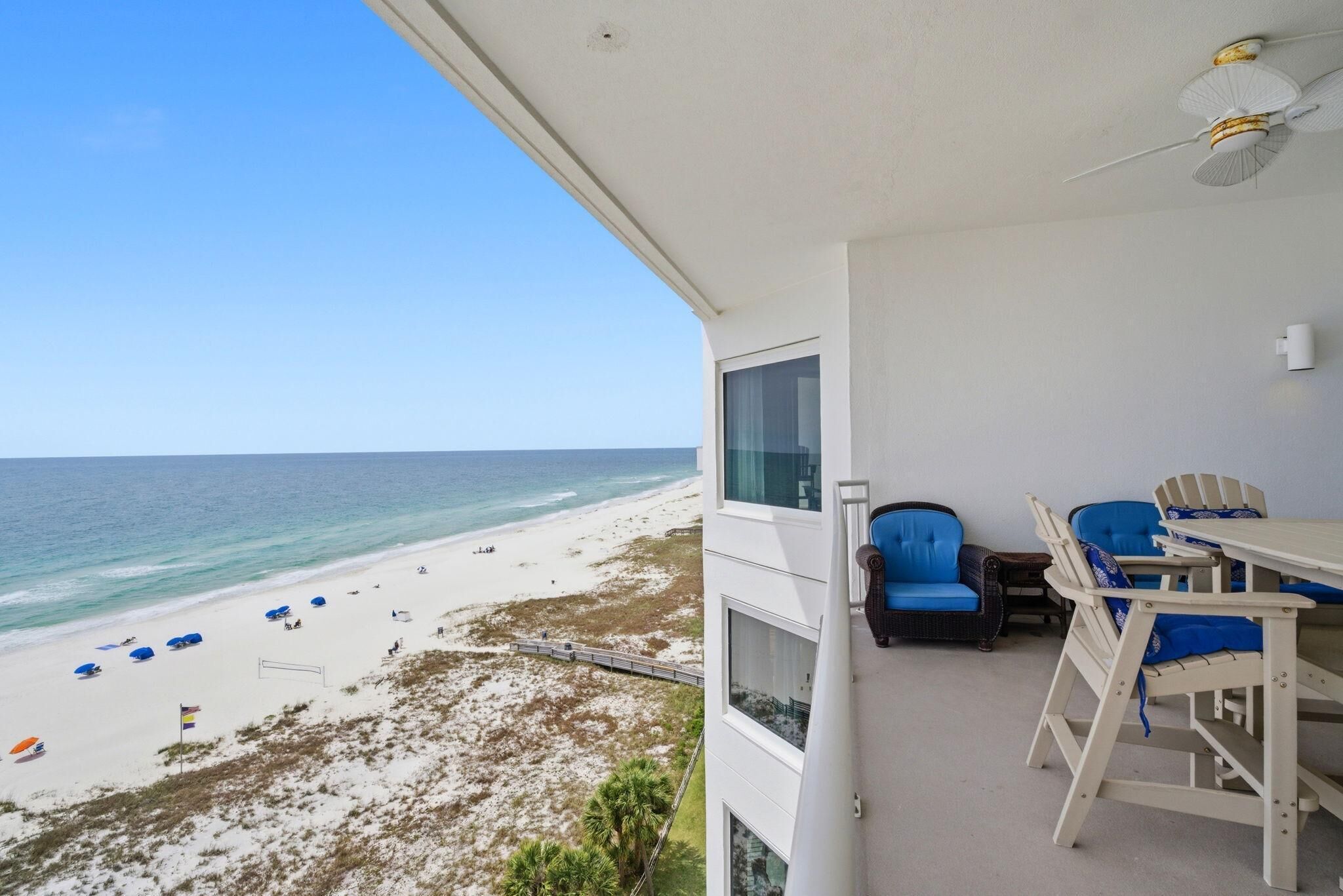 A balcony with a view of a beach, a table with chairs, and a ceiling fan.