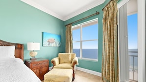 A bedroom with a bed, a chair, a nightstand, a lamp, a painting, and a large window with curtains. - Seaspray East 1023- Gulf Front (Pensacola)