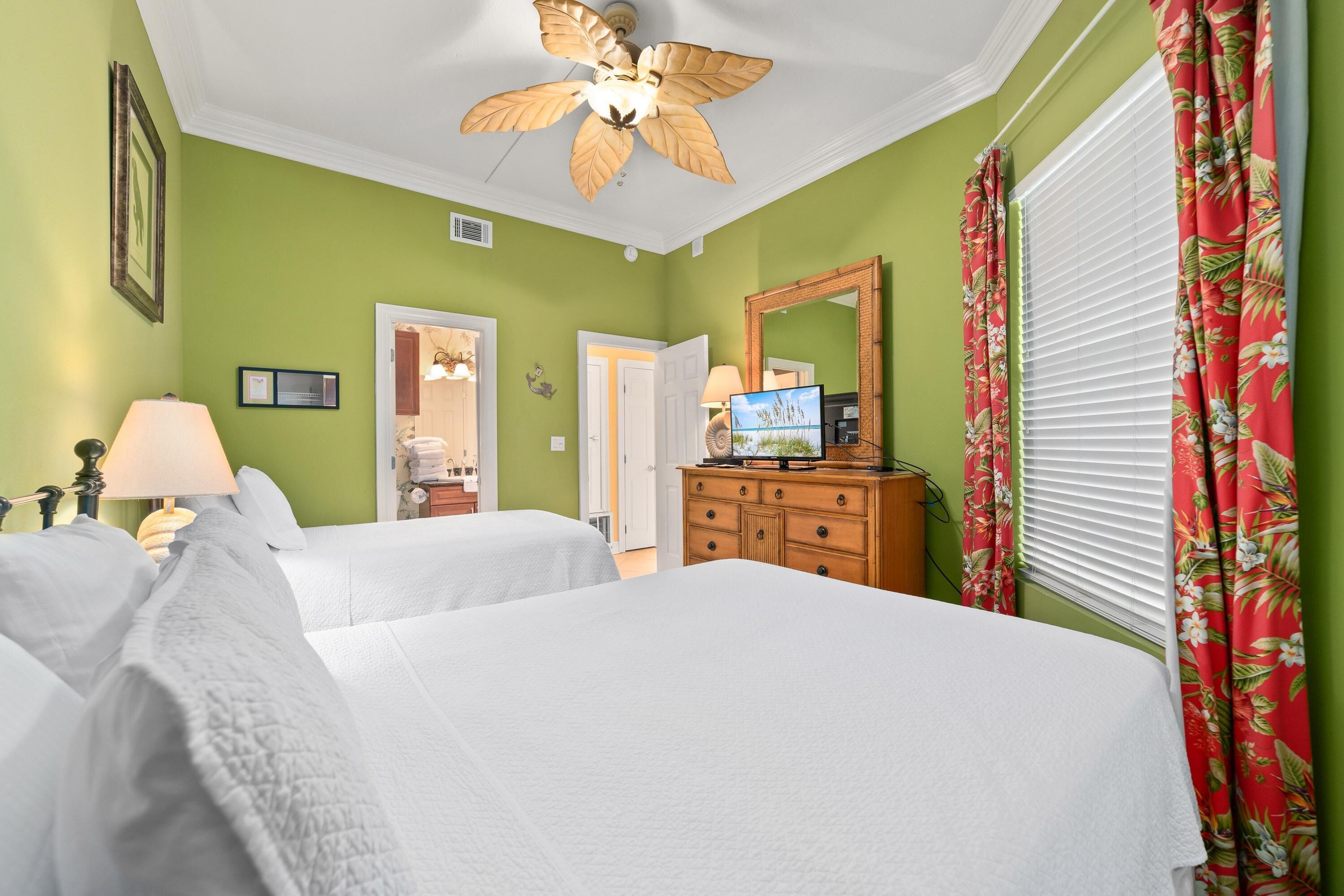 A hotel room with two beds, a wooden dresser, a ceiling fan, a television, and a window with curtains.