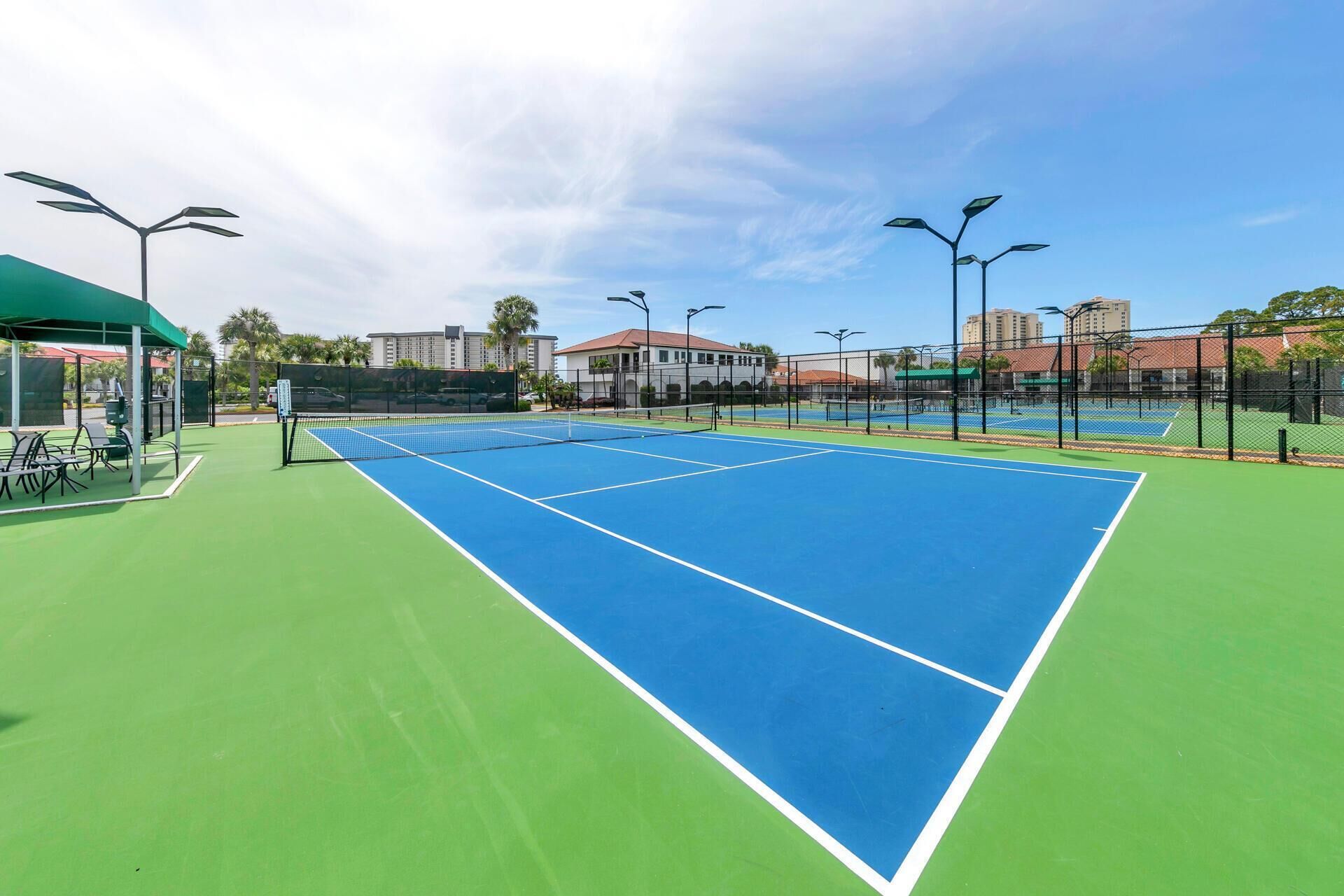 En tennisbana med gröna och blå ytor, omgiven av ett staket, och ett takförsett sittområde med stolar.