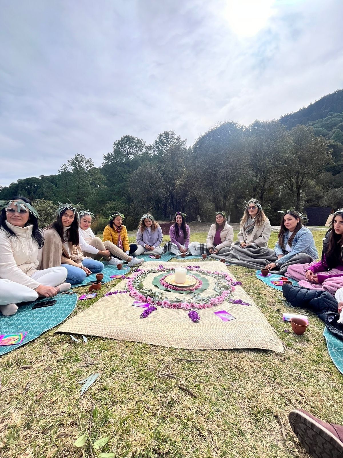 A group of people sitting on mats outdoors with a decorated circle in the center, surrounded by trees and hills.