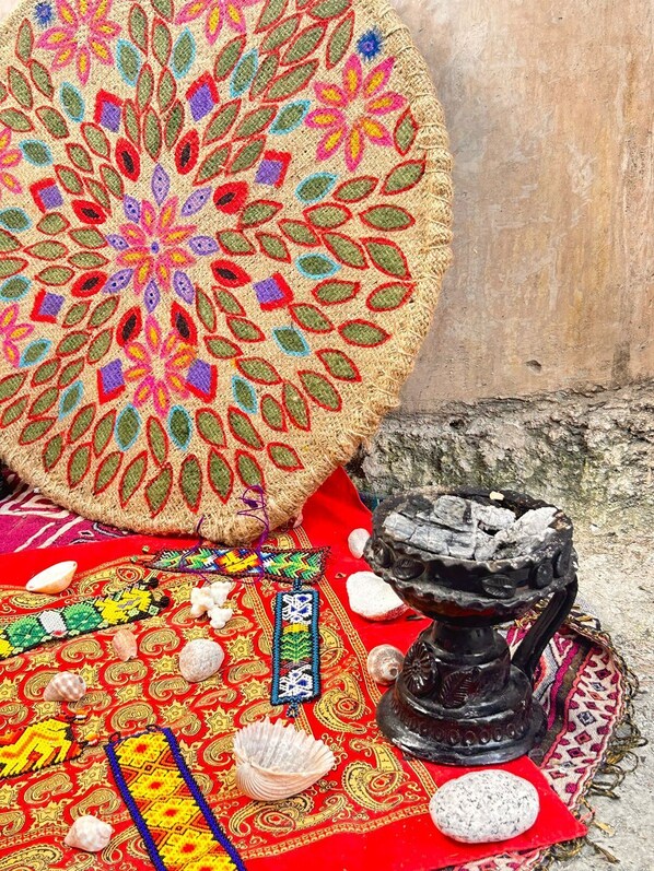 A traditional woven mat with a colorful floral pattern, a small black stone altar, and various shells and beads on a red patterned cloth.