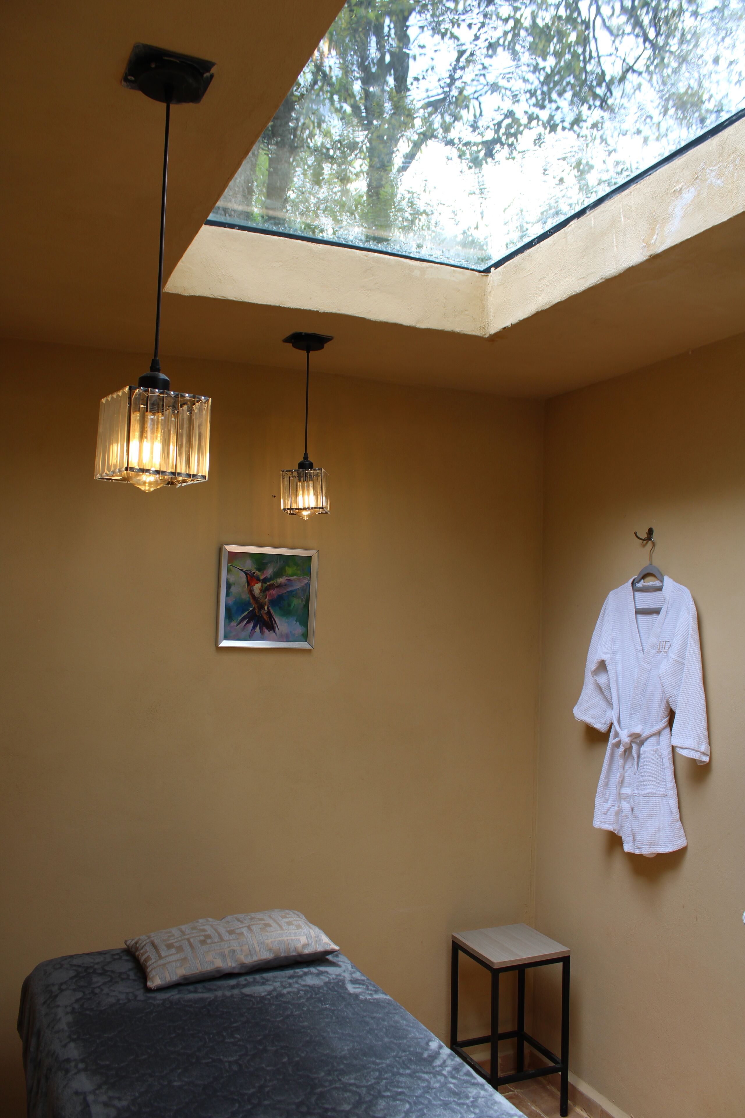 A room with a bed, a small table, a hanging robe, and a painting.