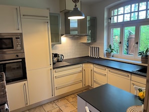 A kitchen with white cabinets, a microwave, a kettle, and a window with a view of greenery.