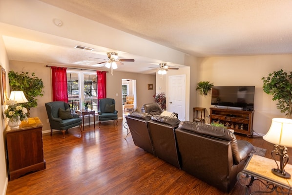A spacious living room with hardwood floors, a leather sofa, a green armchair, a dining table, and a ceiling fan.