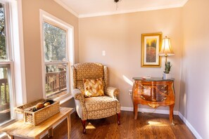 A cozy room with a patterned armchair, a wooden side table with a lamp, and a window with a view of trees.