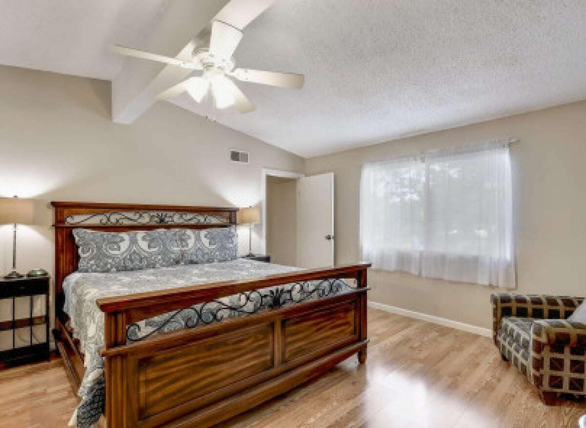 Un dormitorio con cama de madera, ventilador de techo, mesita de noche, una silla y ventana con cortinas.