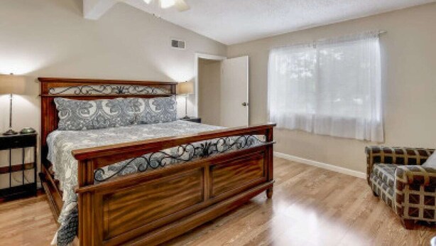 Une chambre à coucher avec un lit en bois, un ventilateur de plafond, une table de chevet, une chaise et une fenêtre avec des rideaux.