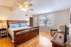 A bedroom with a wooden bed, a ceiling fan, a window with blinds, a television on a stand, and a chair.