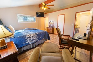 A bedroom with a bed, a desk, a chair, a ceiling fan, a TV, and a window with blinds.