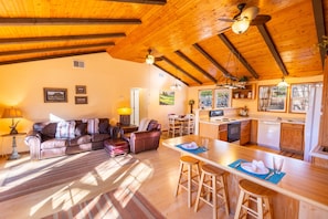 A spacious living room with a wooden ceiling, a dining area with a kitchen in the background, and a large window providing natural light. - Wimberley Getaway with Private Pool and Fire Pit (Wimberley)